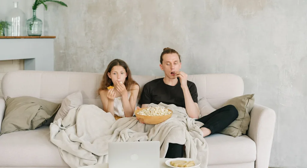 Stock photograph: two people sitting on a pale sofa under a blanket with a bowl of popcorn, watching a laptop on the table in front of them
